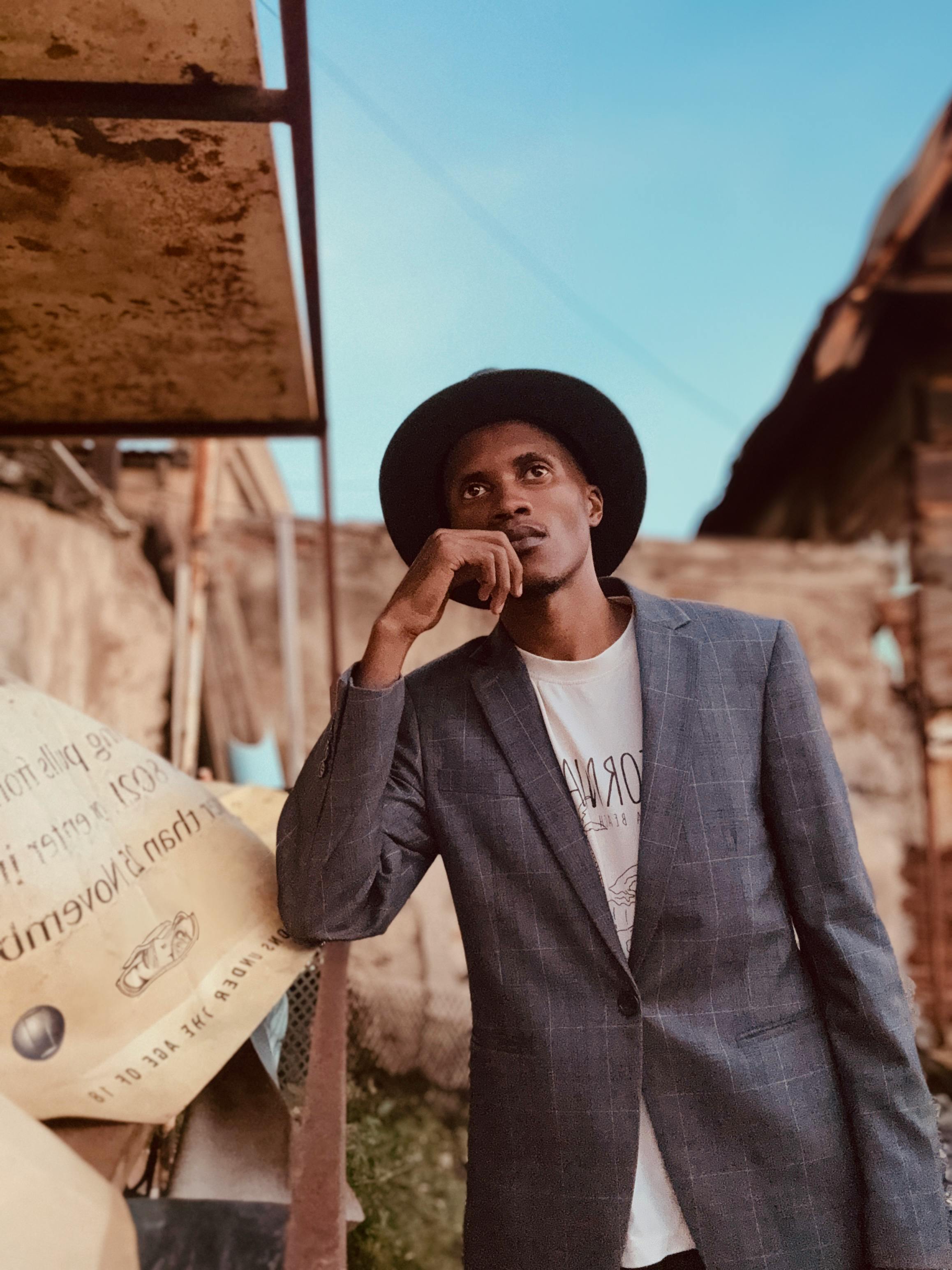 A stylish young man in a suit and hat ponders thoughtfully in an urban environment.