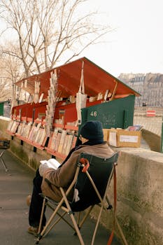 A man enjoys reading by a classic Parisian book stall along the Seine River.