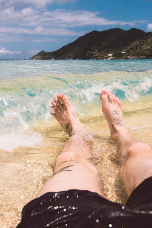 문신을 한 다리와 해안으로 튀는 파도와 함께 해변에서 Lazing하는 사람의 발의 근접
