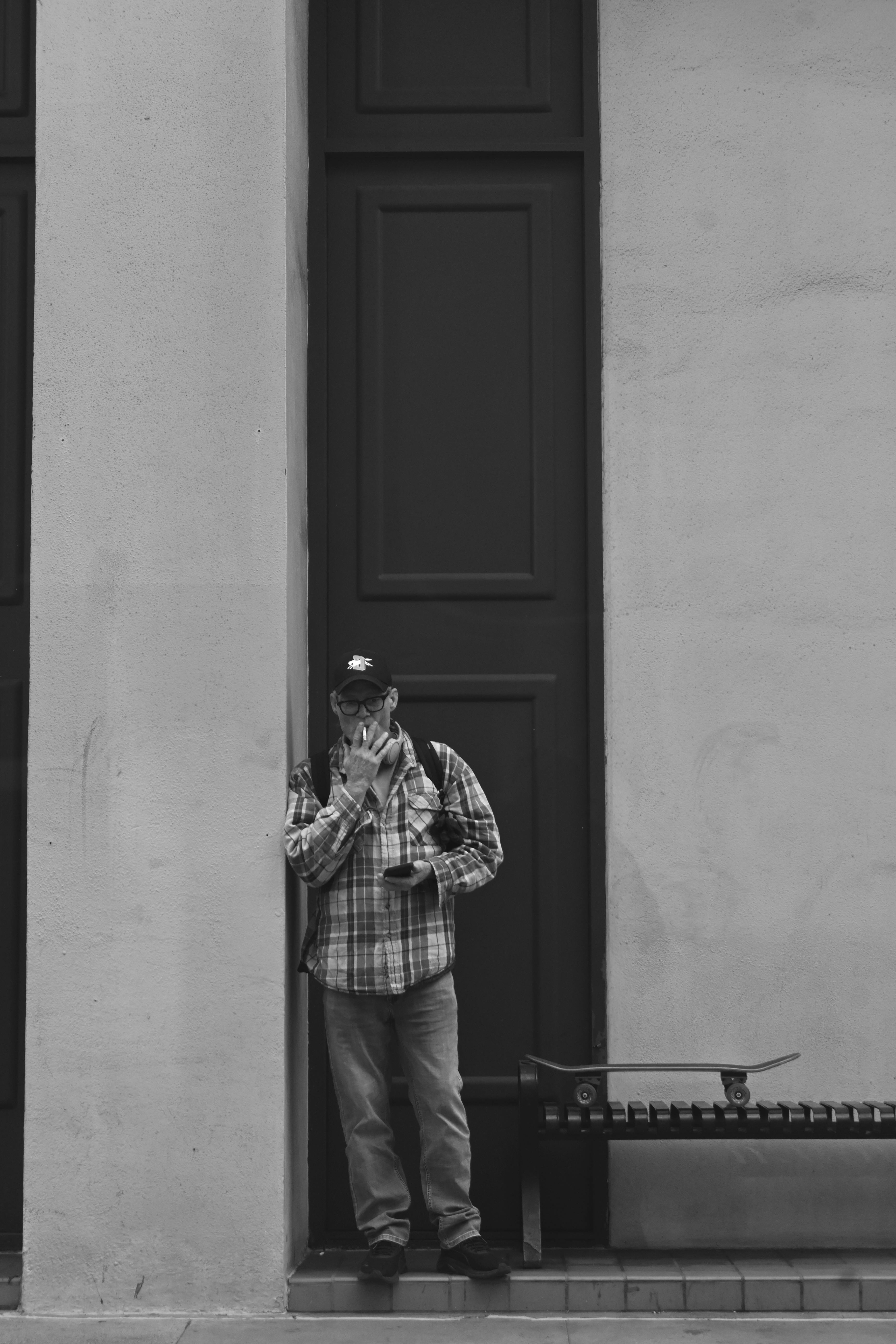 Gratuit Photo de rue en noir et blanc d'un homme fumant près d'un skateboard. Photos