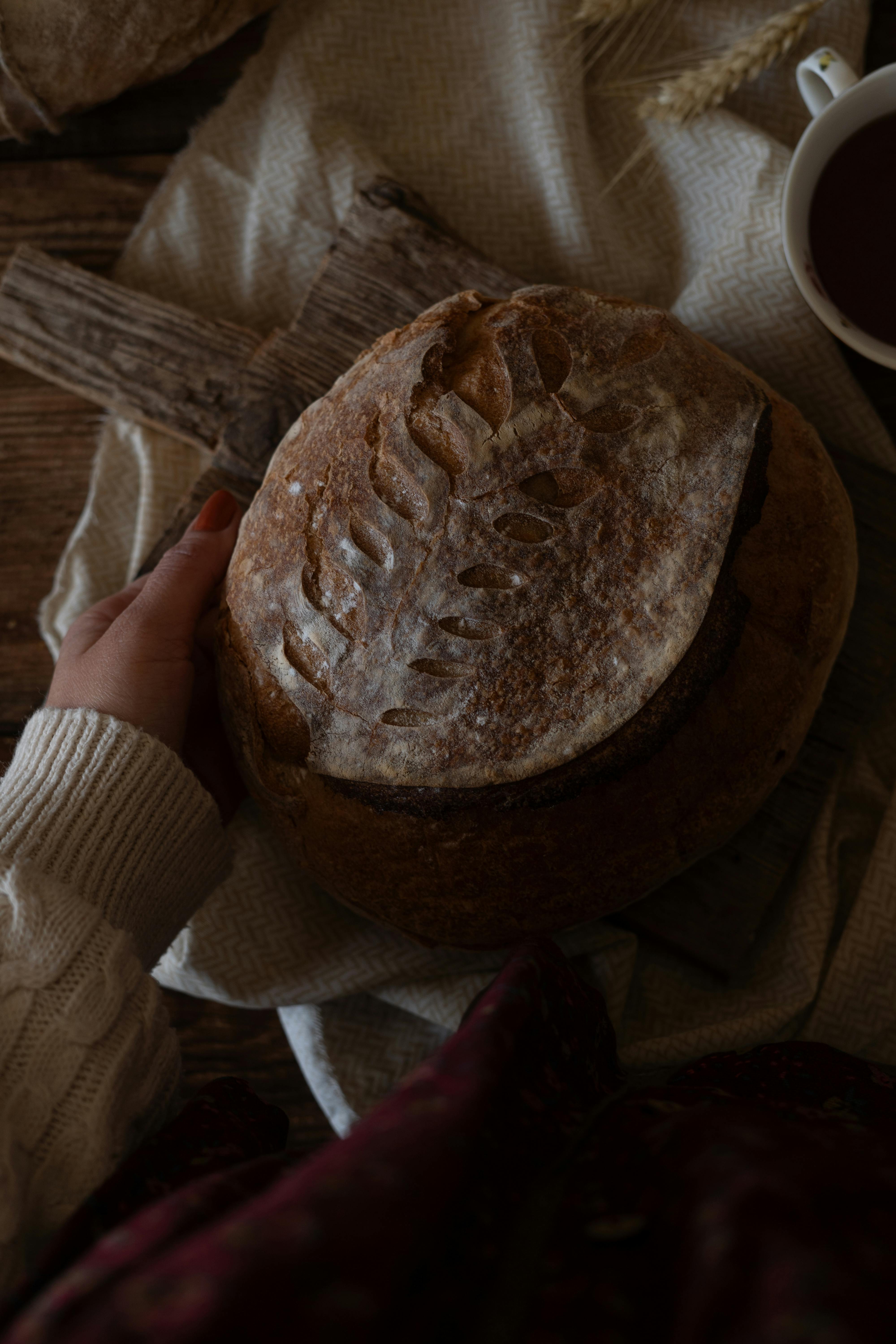 Hand holding rustic artisan bread with intricate leaf pattern and warm textile.