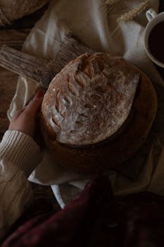 Hand holding rustic artisan bread with intricate leaf pattern and warm textile.