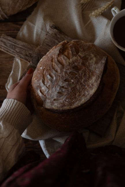 Hand holding rustic artisan bread with intricate leaf pattern and warm textile.