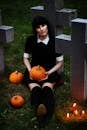 一位哥特风格的女子坐在墓地中，手持南瓜，周围环绕着南瓜与蜡烛，营造出怪异的万圣节氛围