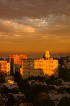 Golden hour cityscape with an airplane, buildings cast long shadows under a vibrant sky.