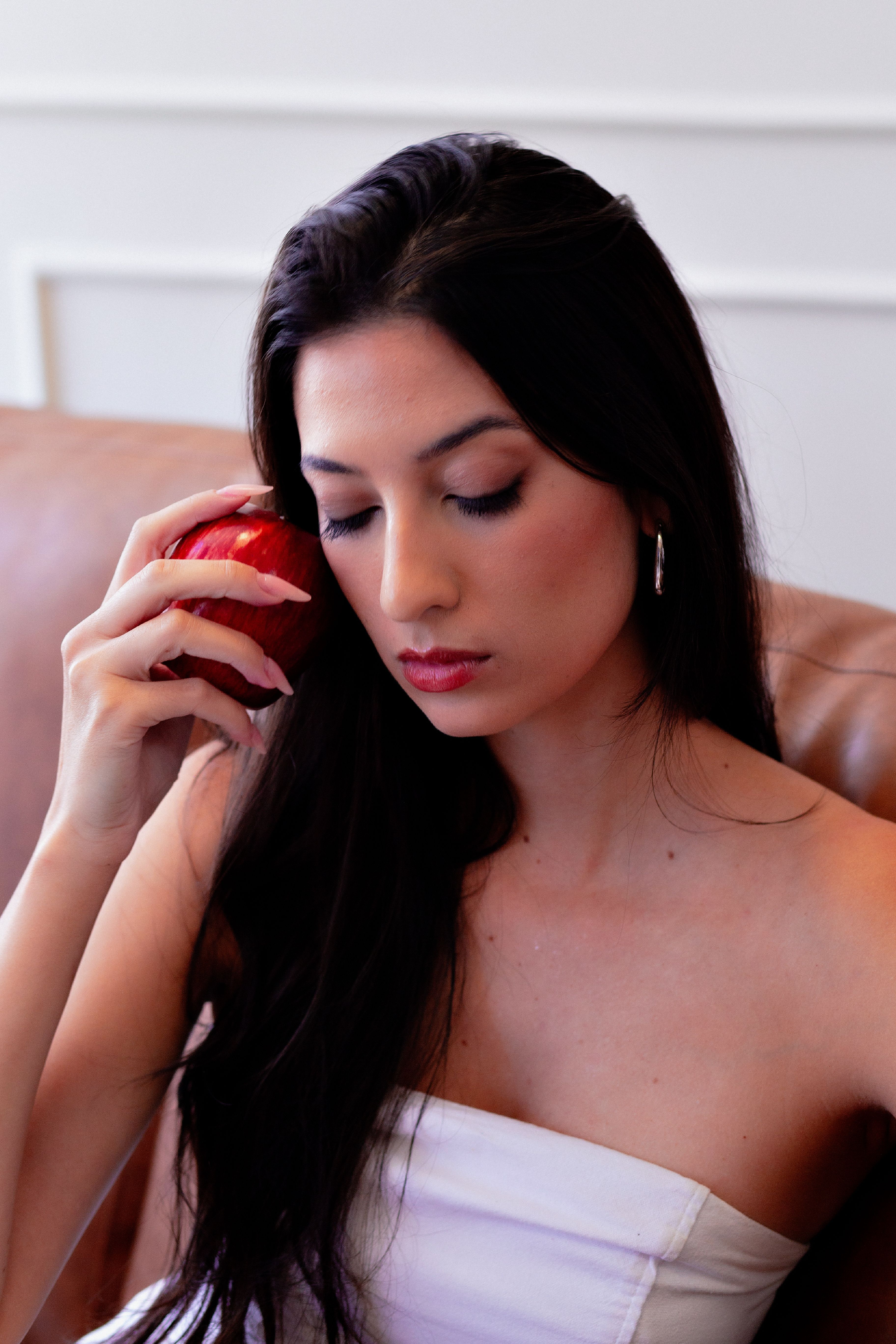 A thoughtful woman with long hair holds a red apple indoors.