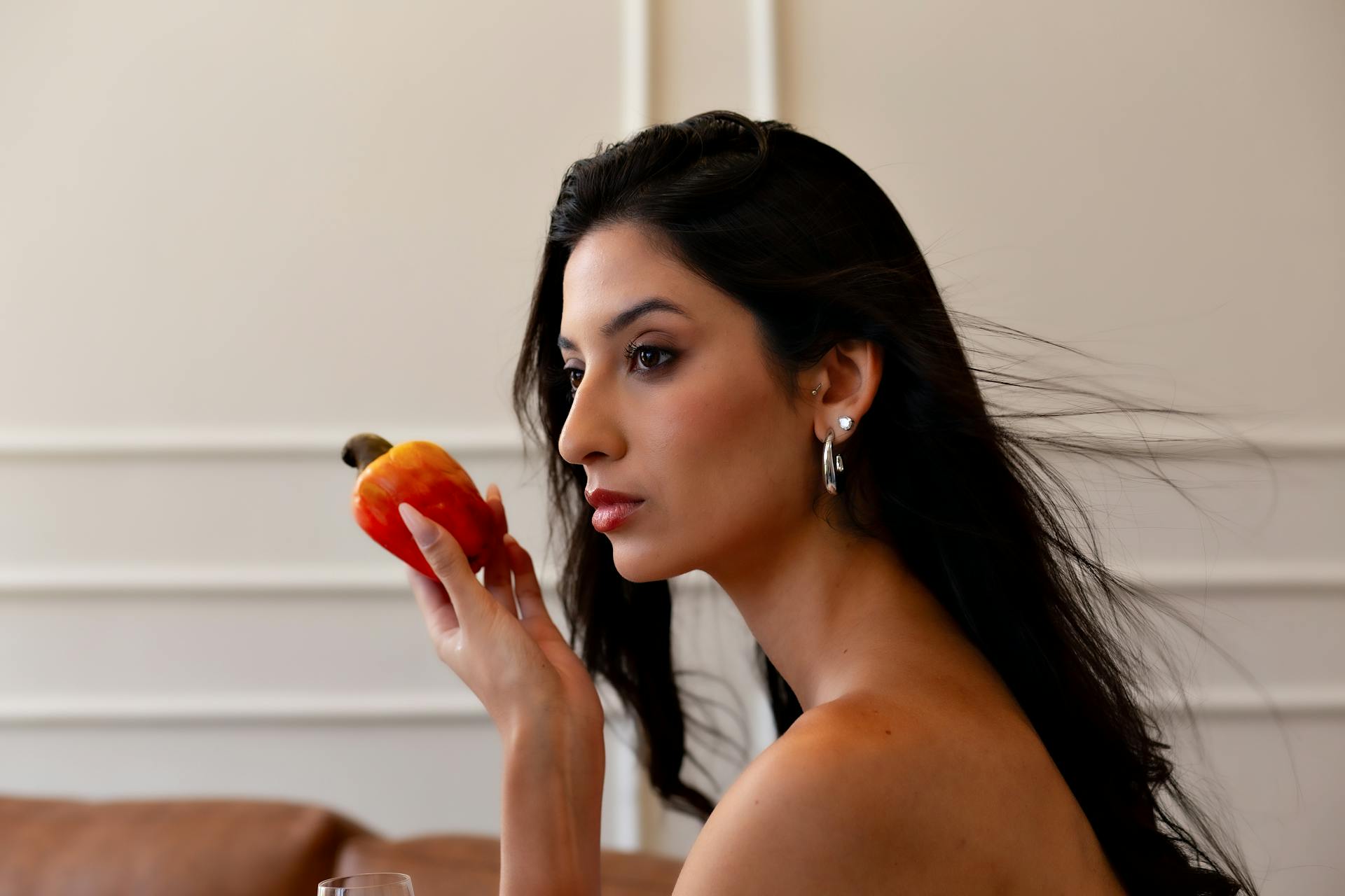 A woman gazes thoughtfully at a vibrant cashew apple in an indoor setting.