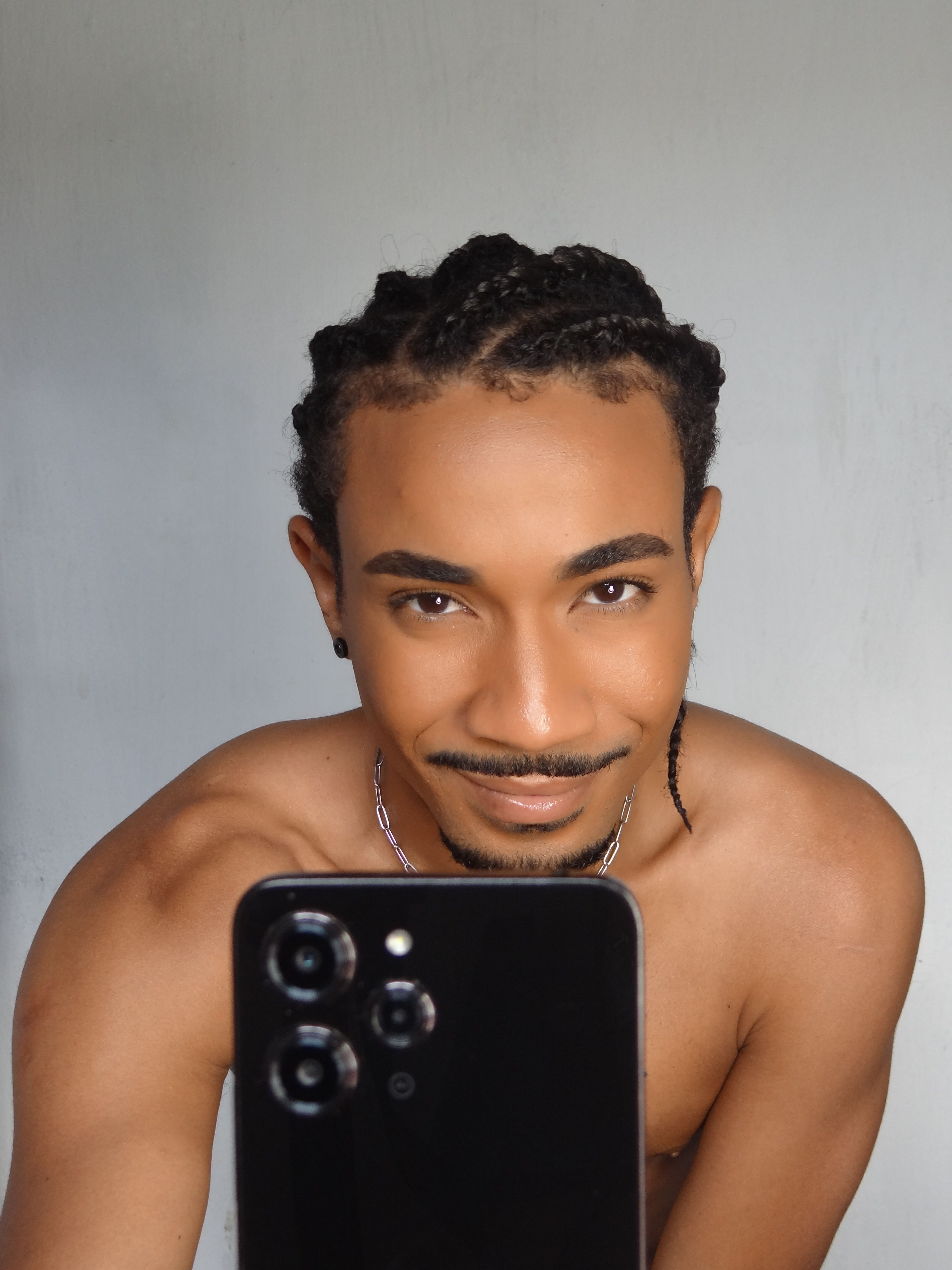 A smiling man takes a selfie indoors, showcasing his braided hairstyle.