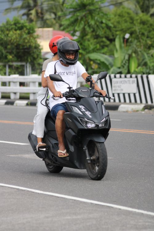 Free Two adults on a scooter driving on a road, surrounded by greenery. Safety gear included. Stock Photo