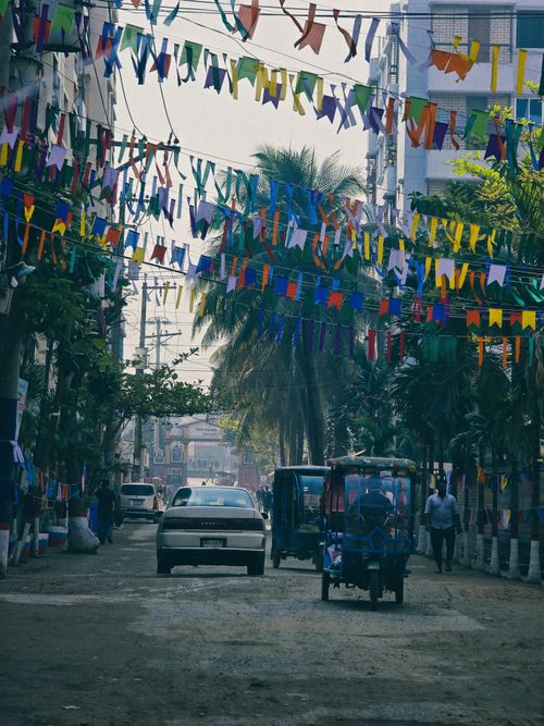 Free A lively street in Dhaka adorned with colorful flags and bustling with vehicles and pedestrians. Stock Photo