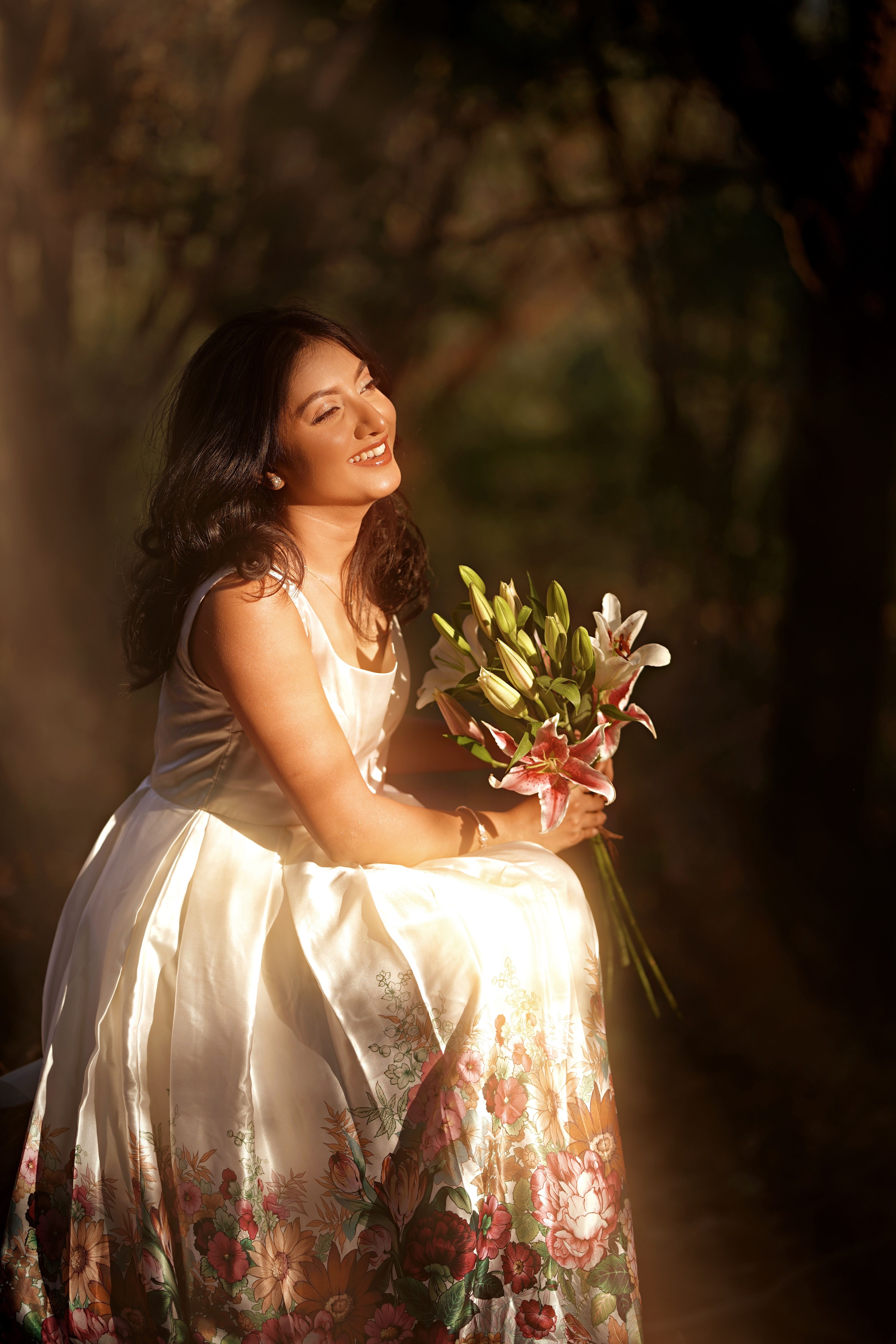 A woman in a floral dress smiles while holding lilies in a sunlit forest.