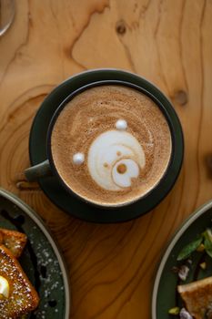 A charming bear design in latte art above a rustic, cozy breakfast setting.