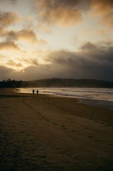 A serene sunset scene on a beach in Armação dos Búzios, RJ, Brazil, with two people strolling along the shoreline.