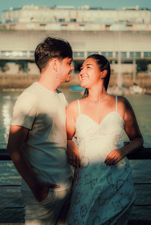 Free A couple smiles warmly at each other by the water in sunlight. Stock Photo