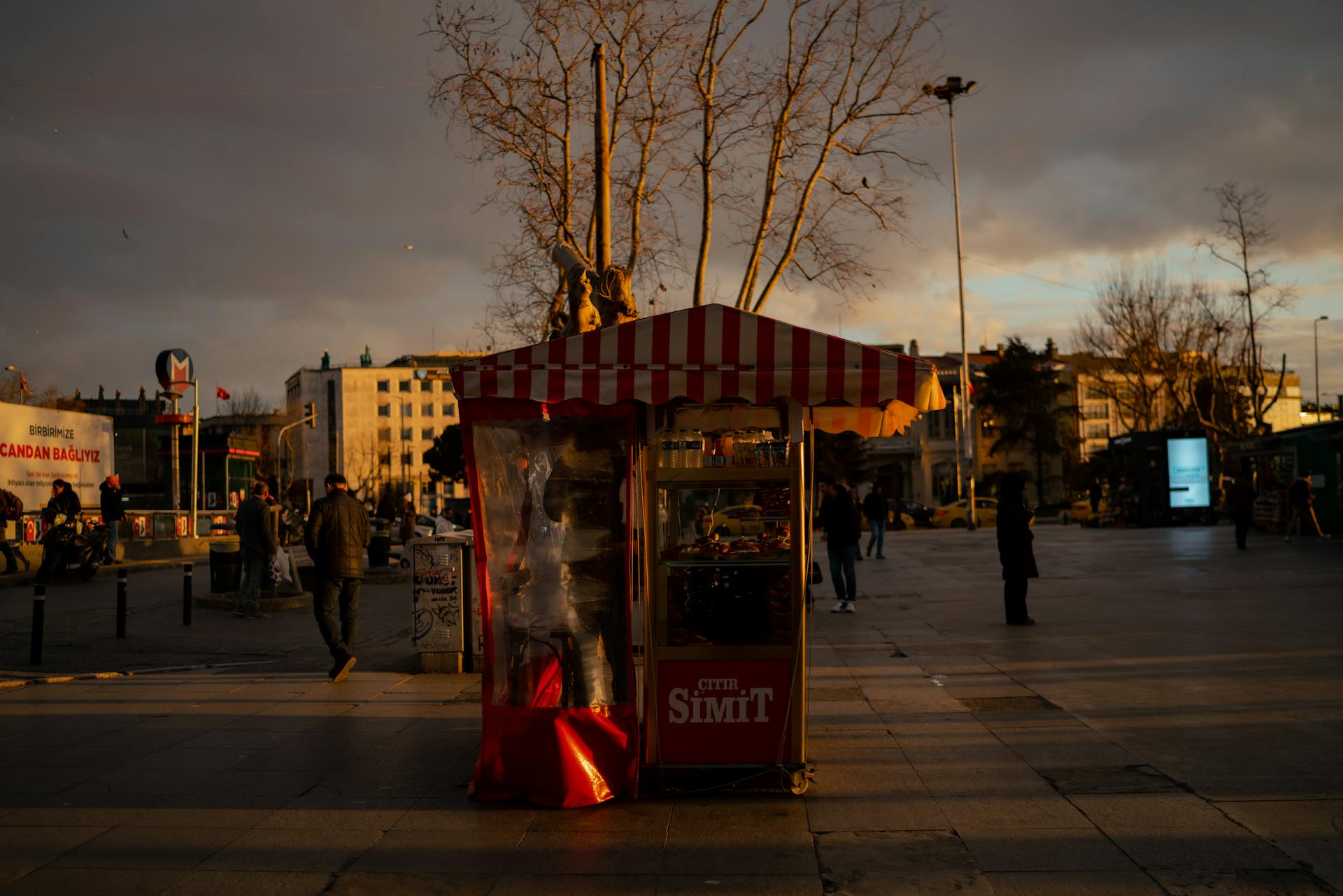 A simit vendor stall in a bustling urban plaza at sunset, casting long shadows.