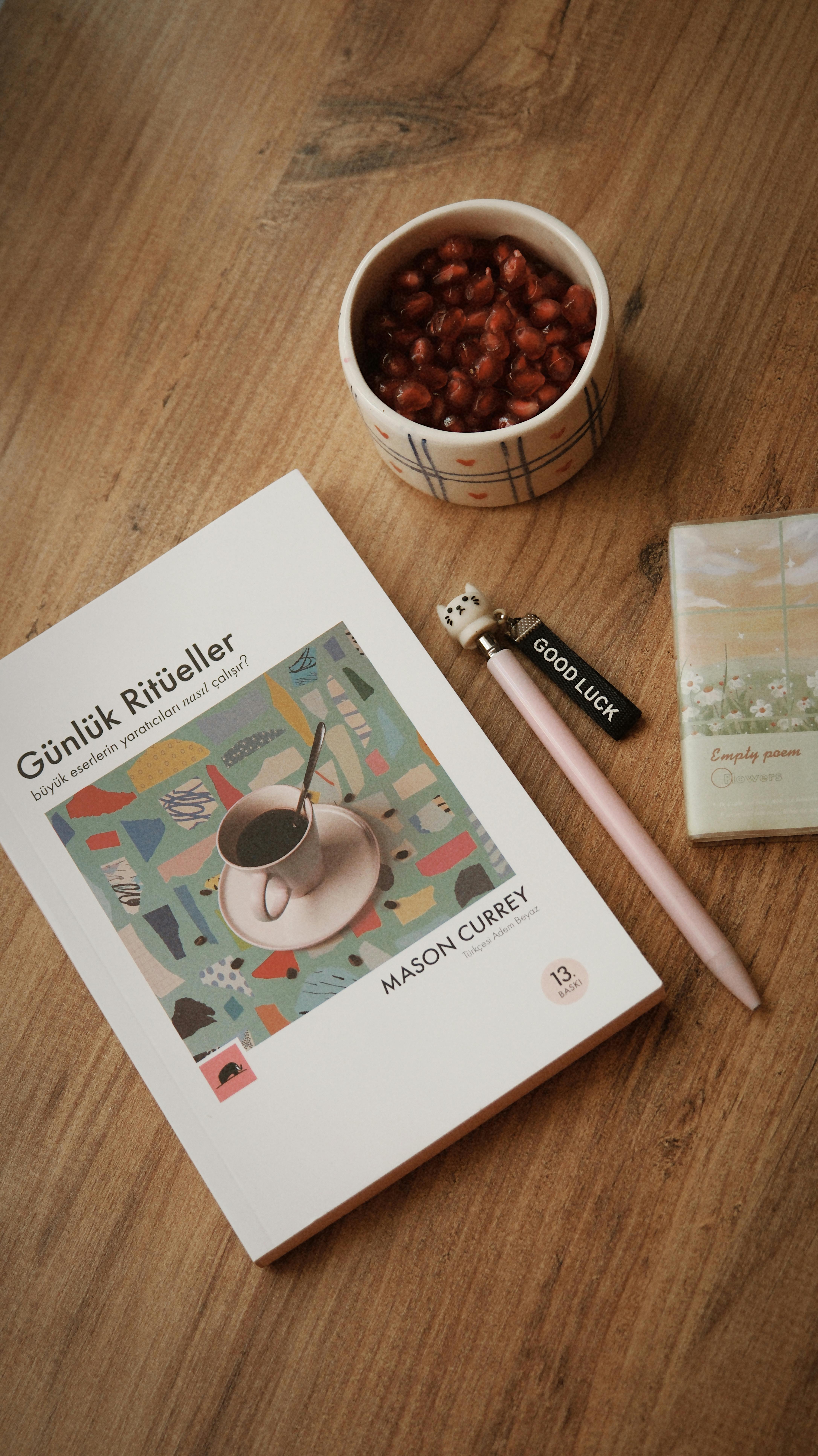 Warm flat lay of a book, pomegranate seeds, and a cute pen on a wooden table.