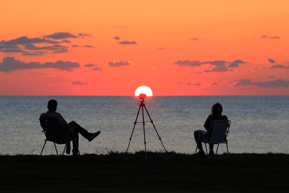 Casal sentado à beira-mar, em silhueta, diante do pôr do sol