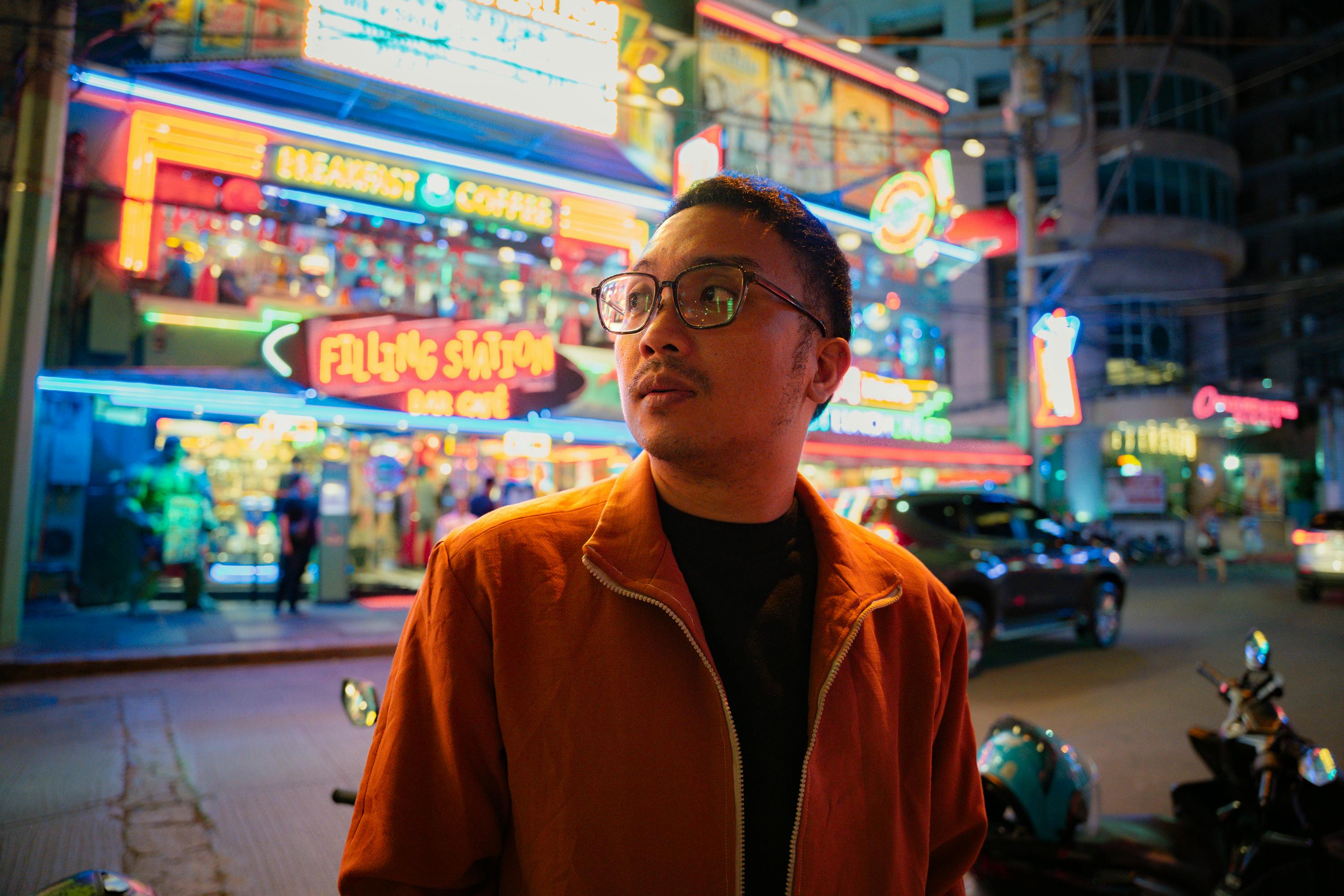 Free Man in an orange jacket stands in a colorful city street at night, with neon signs and bustling nightlife. Stock Photo