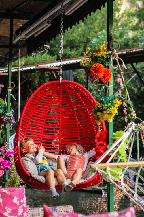 Free Two kids enjoying a colorful red hanging chair outdoors surrounded by vibrant flowers. Stock Photo
