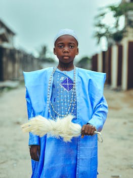 Young boy dressed in vibrant blue traditional clothing stands outdoors holding a white feather accessory.