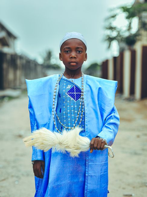 Young boy dressed in vibrant blue traditional clothing stands outdoors holding a white feather accessory.