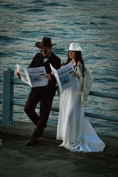 A fashionable couple in stylish hats reading newspapers by the waterfront.