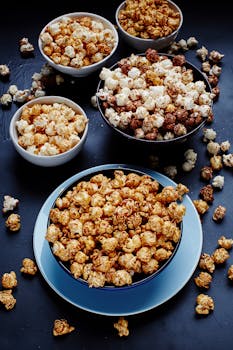Top view of assorted flavored popcorn in bowls, showcasing sweet and salty varieties on a dark surface.