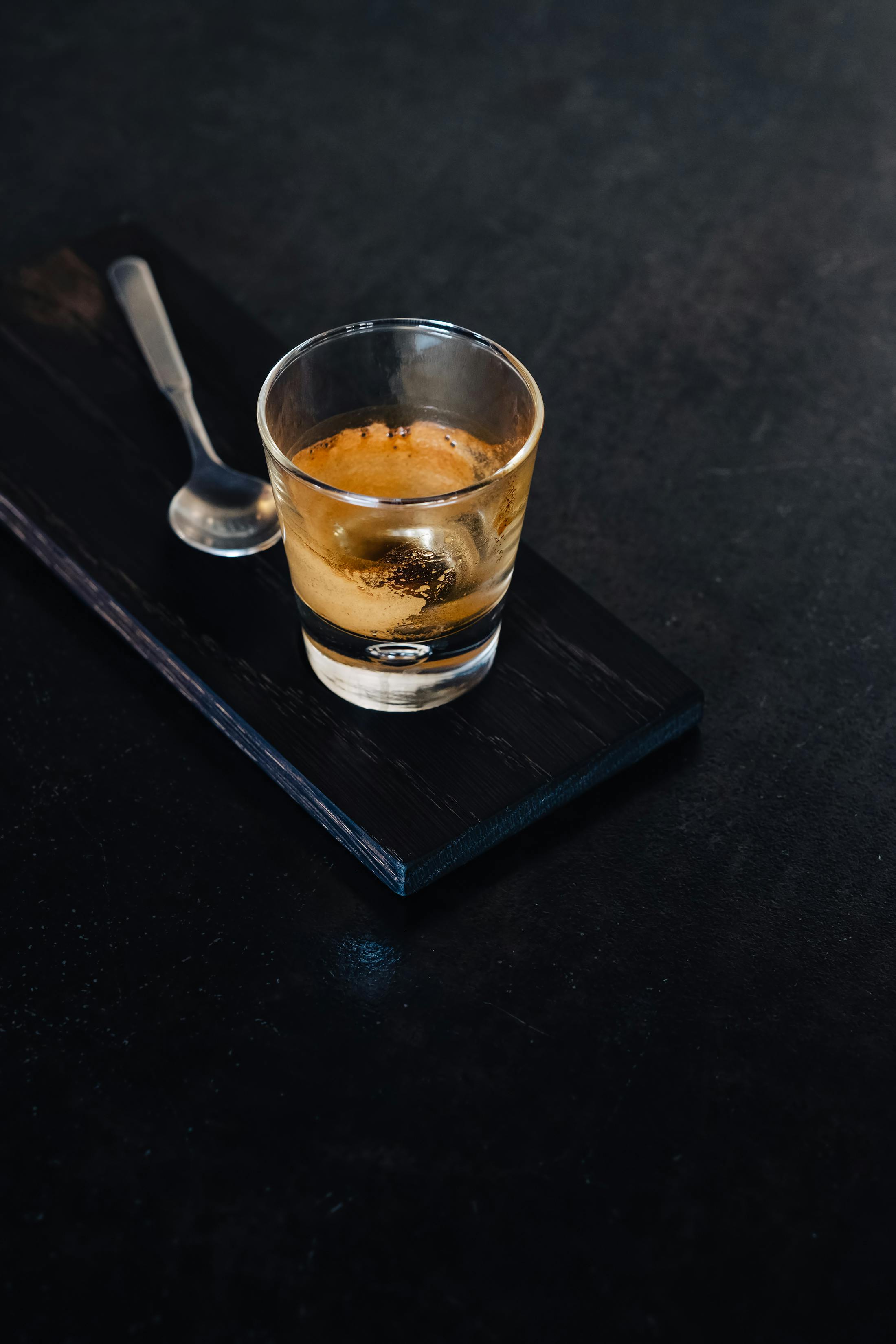 A minimalist image of espresso served in a glass with a spoon on a dark wooden board.