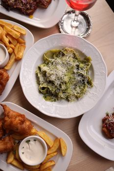 Overhead shot of creamy pesto pasta with cheese and side dishes like fries and fish.