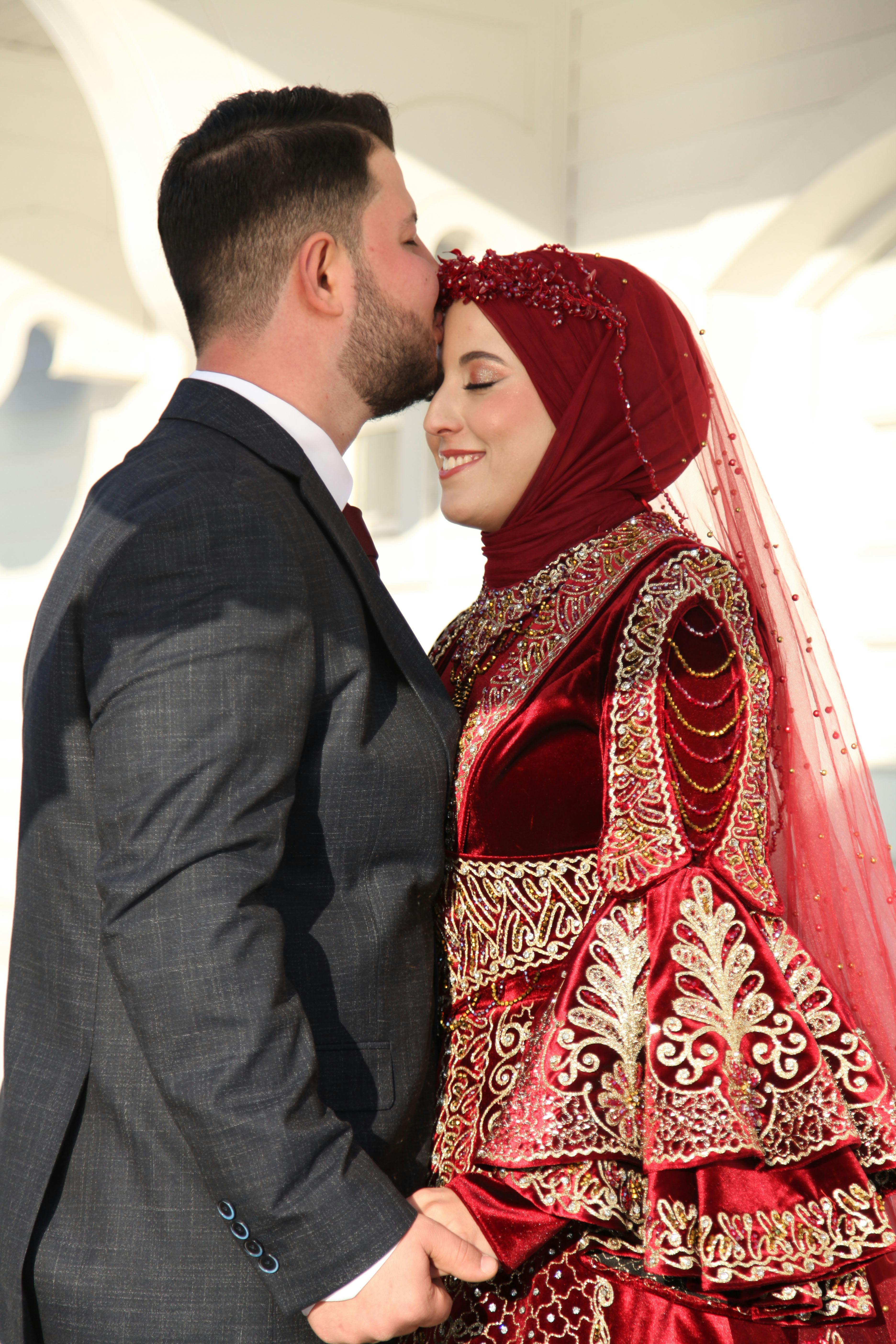 Happy couple embraces in traditional, ornate wedding attire under bright daylight.