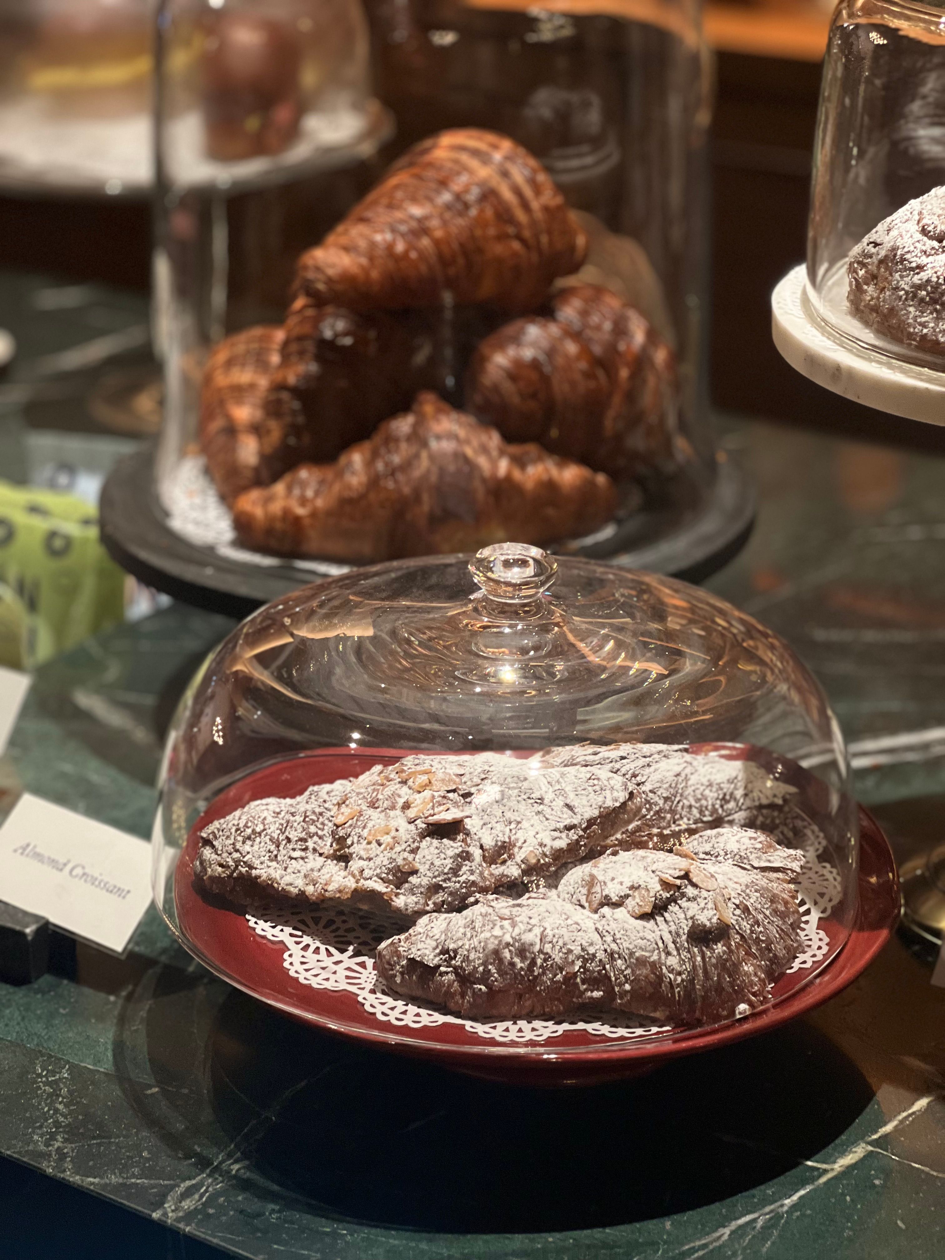 A close-up view of almond and chocolate croissants under glass domes on a dark wooden counter.
