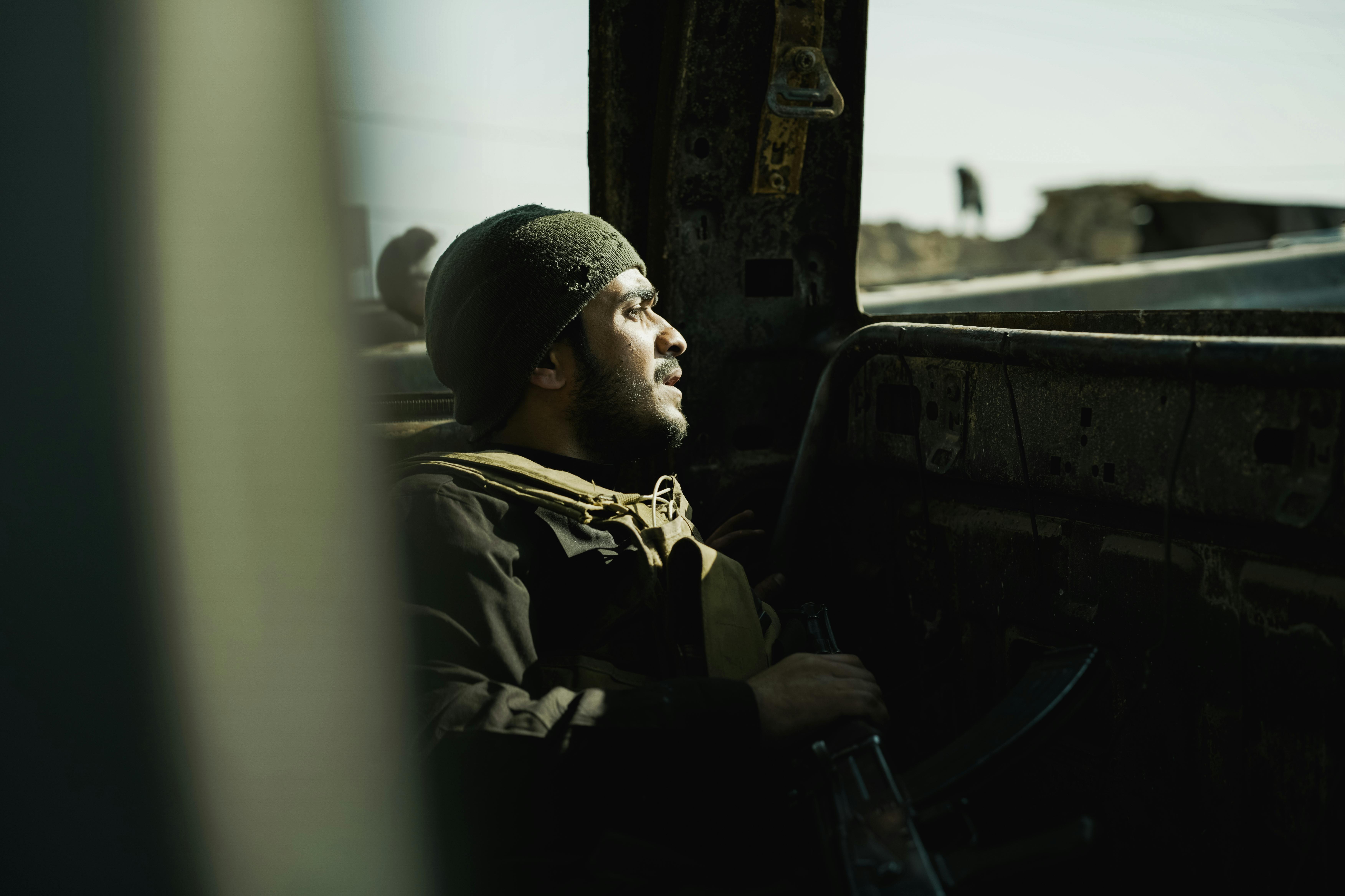 A soldier sits inside a dilapidated structure, deep in thought, illuminated by natural light.