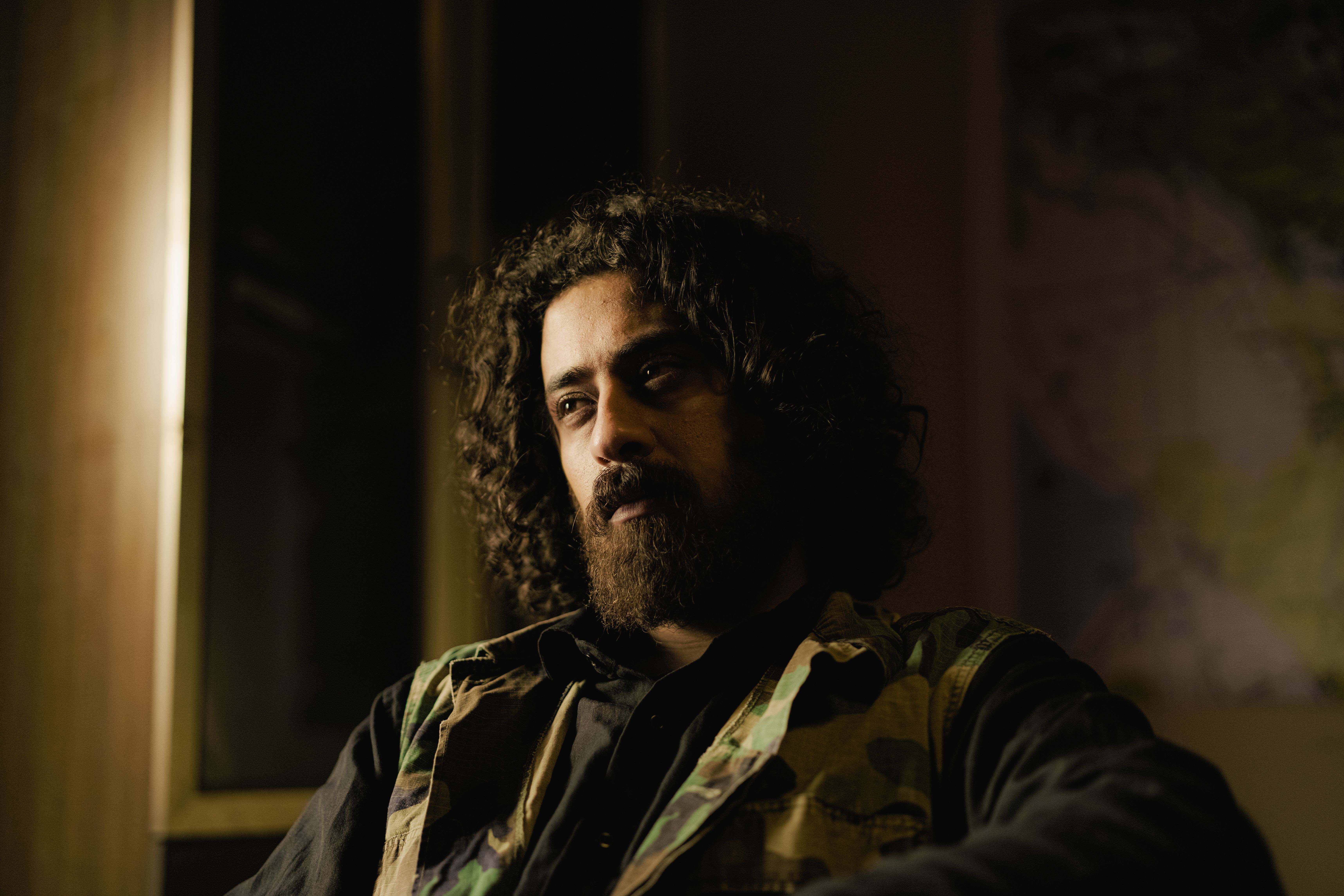 Thoughtful man with curly hair and beard sitting in dimly lit room, wearing dark attire.