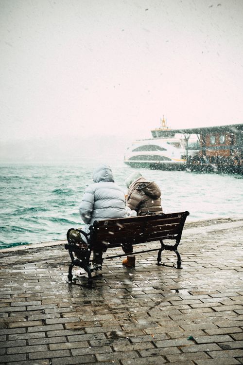 Free A couple in winter coats sits on a bench by a snowy waterfront, watching boats in the distance. Stock Photo