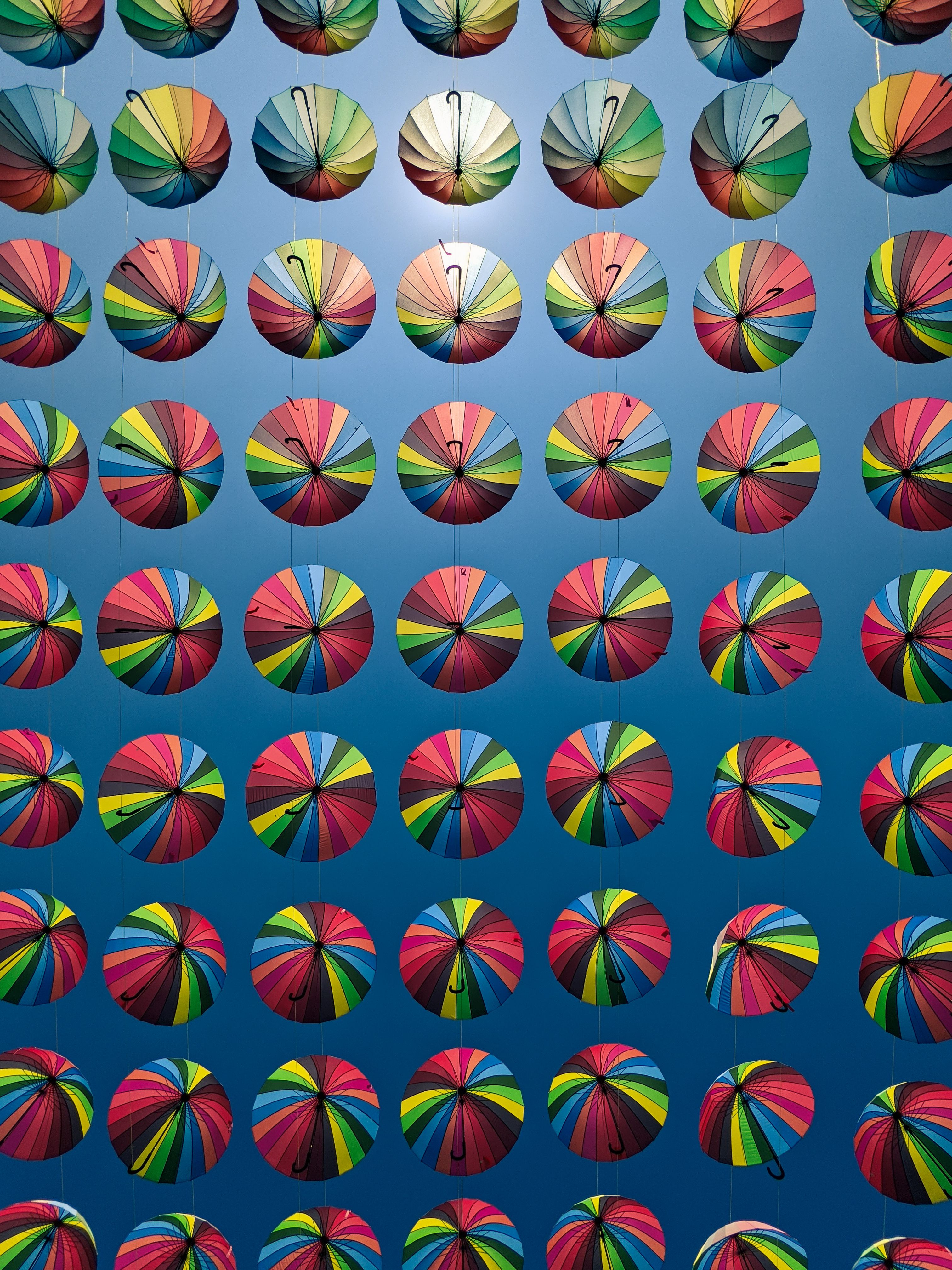 Free Vibrant display of rainbow-colored umbrellas hanging in a geometric pattern against a clear blue sky. Stock Photo