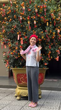 Joyful woman in festive attire poses with decorated kumquat tree during Lunar New Year.