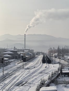 A snow-covered railway leads to an industrial smokestack, emitting smoke against a mountainous backdrop.