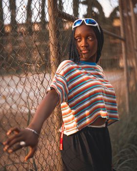Stylish young woman in striped tee and braids leans on a fence, embodying modern urban fashion.