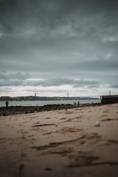 Moody scene of Lisboa's beach with dramatic clouds and cityscape in the distance.