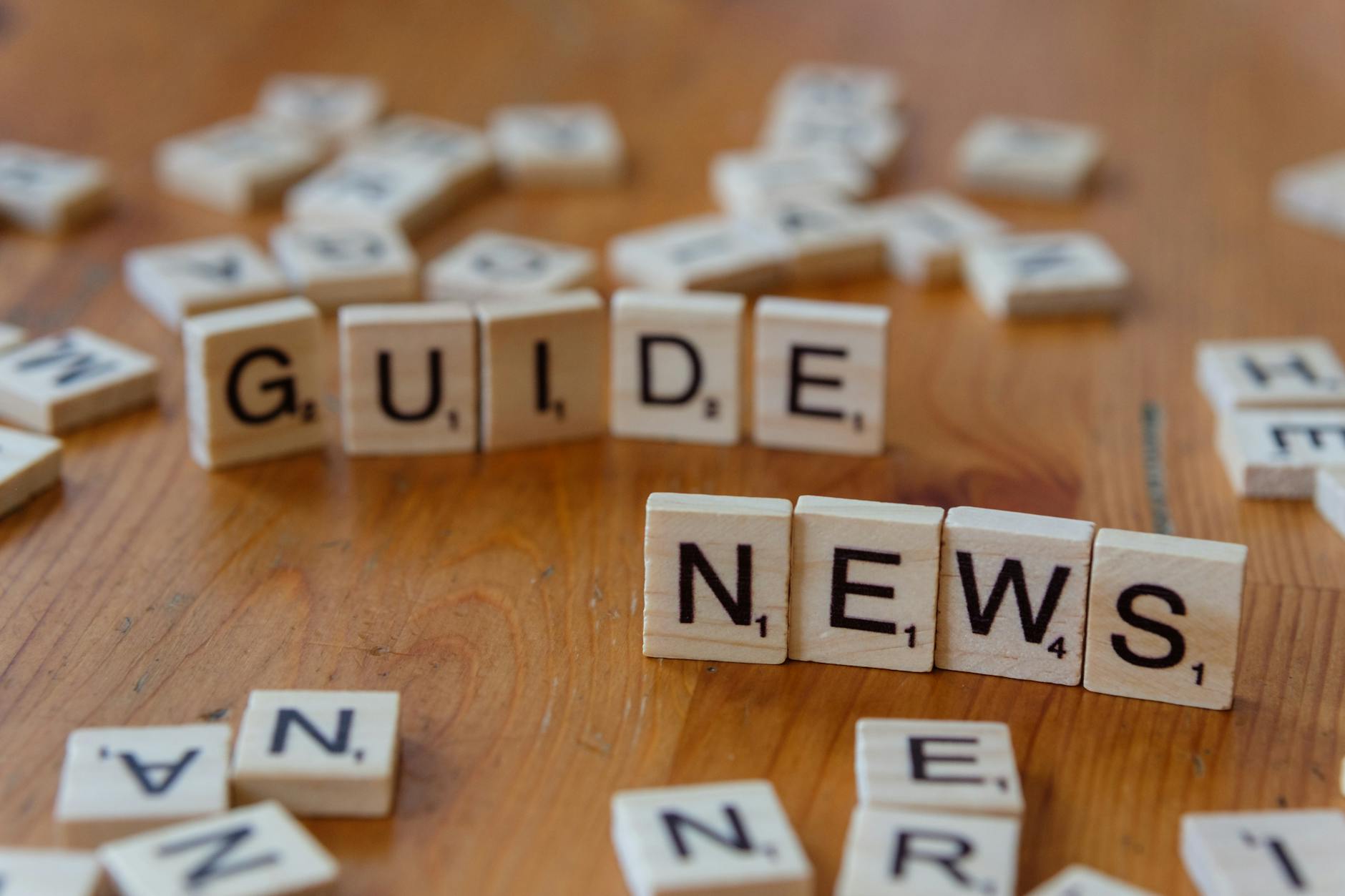 Close-up of Scrabble tiles spelling 'Guide' and 'News' on a wooden surface.