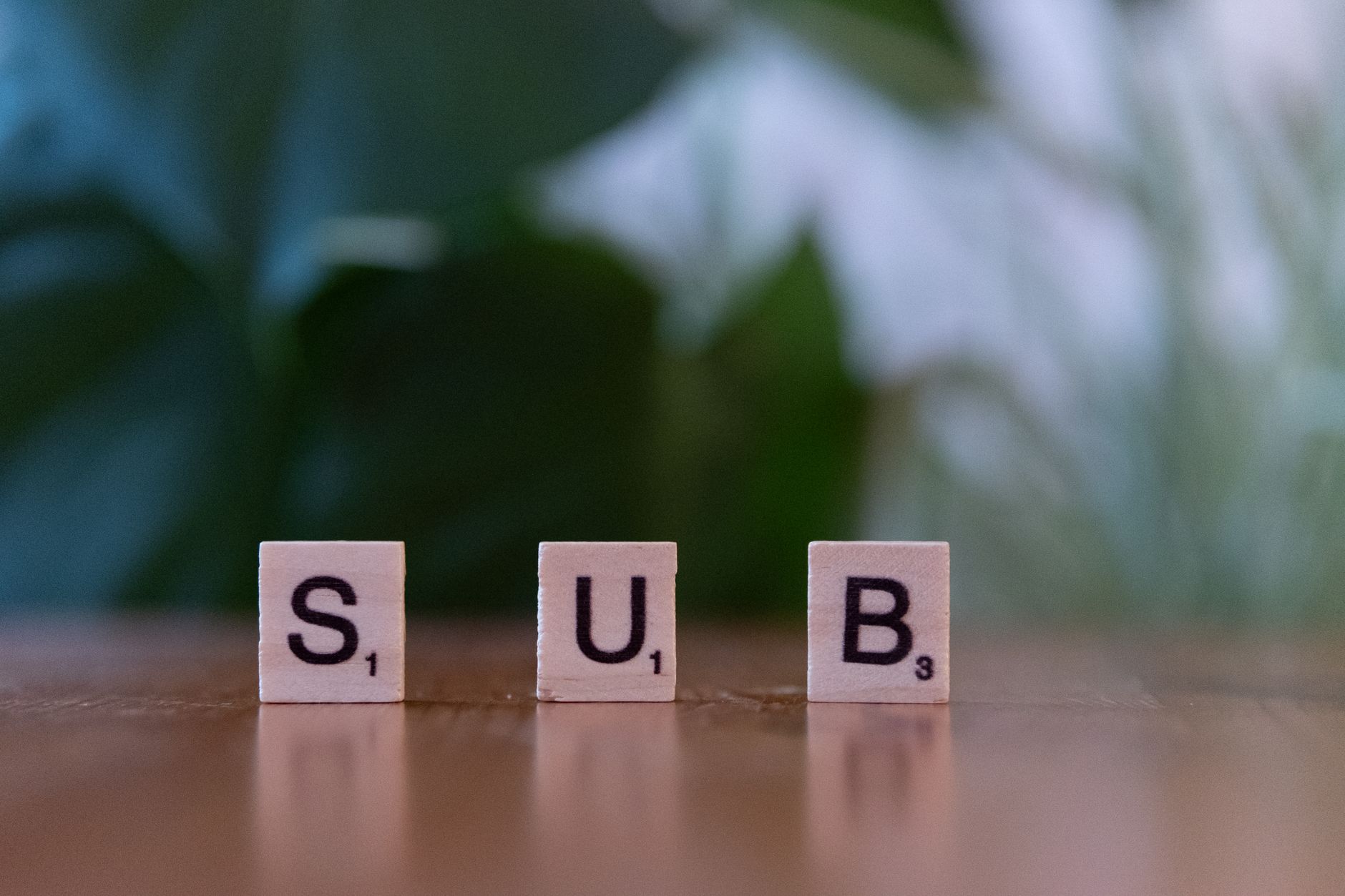 Three Scrabble tiles spelling 'SUB' on a wooden surface with blurred green background.