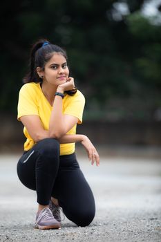 A young woman in a yellow T-shirt kneels outdoors, displaying a confident pose.