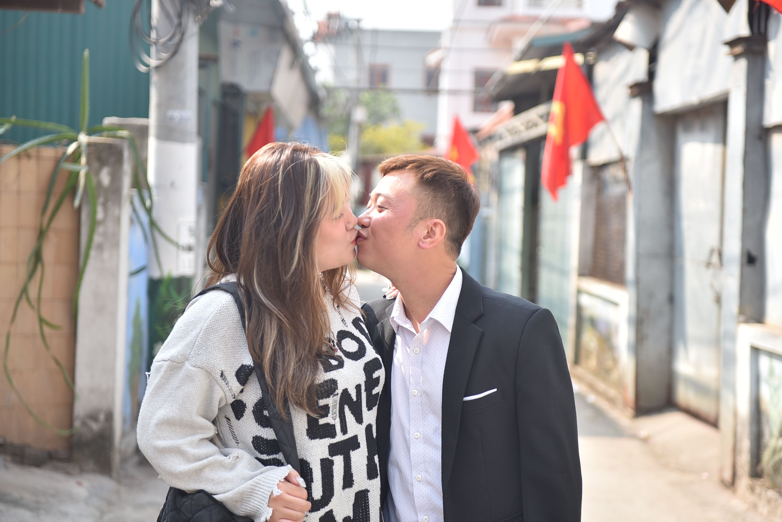 A couple shares a tender kiss on a sunny urban street lined with red flags.