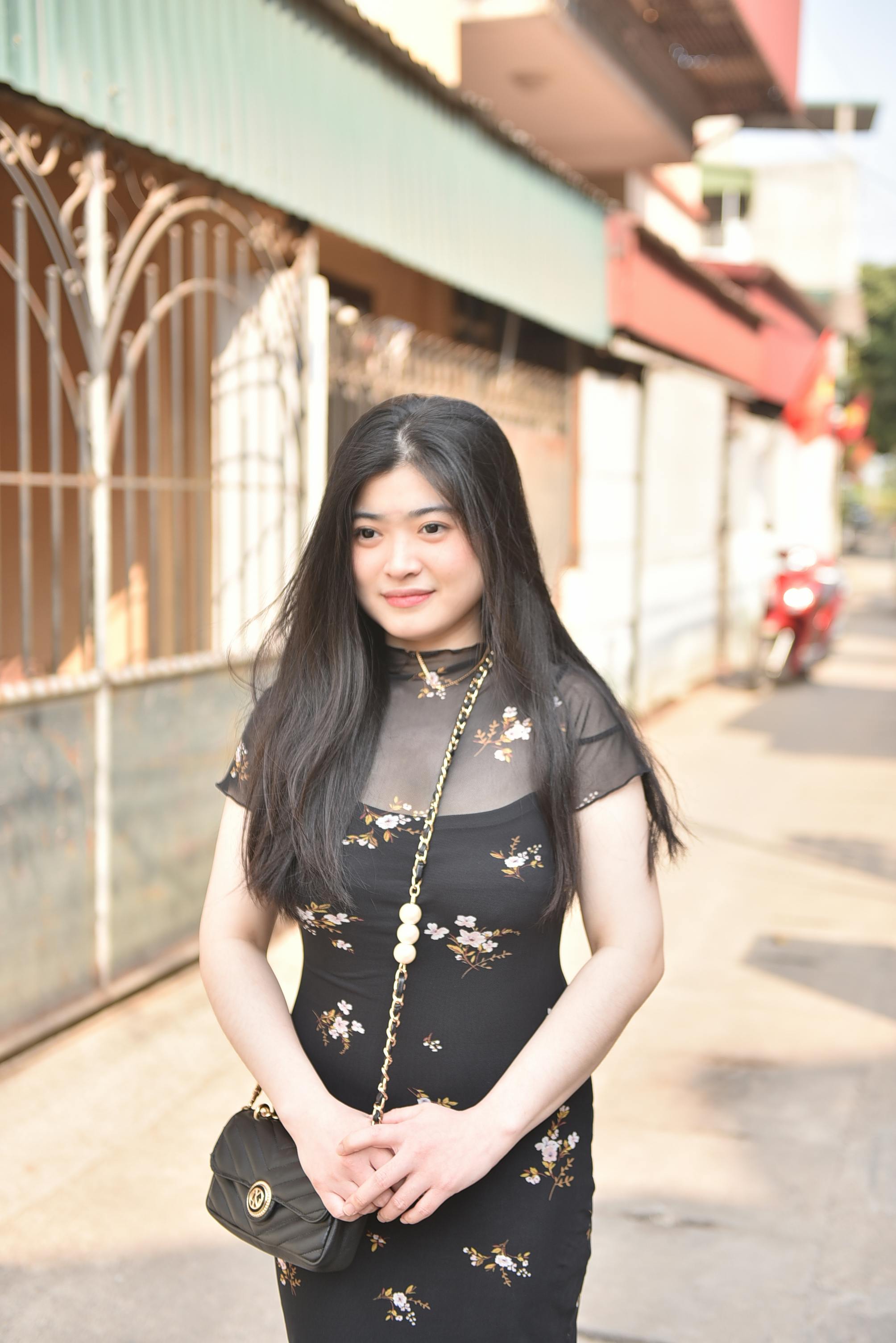 Free A young woman in a floral dress smiles while standing on a sunny city street. Stock Photo