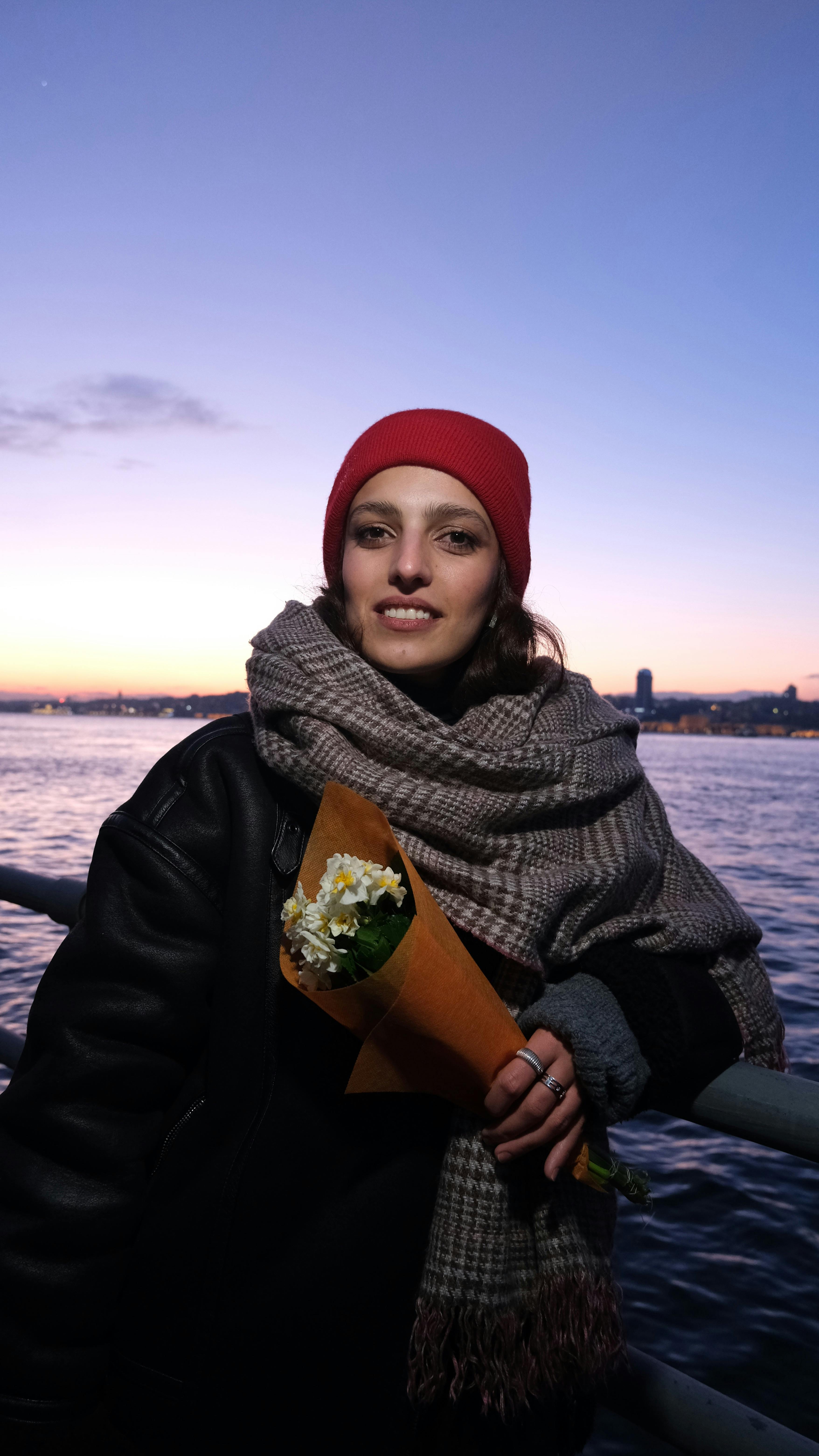 Woman wearing red beanie holding bouquet by waterfront at sunset.