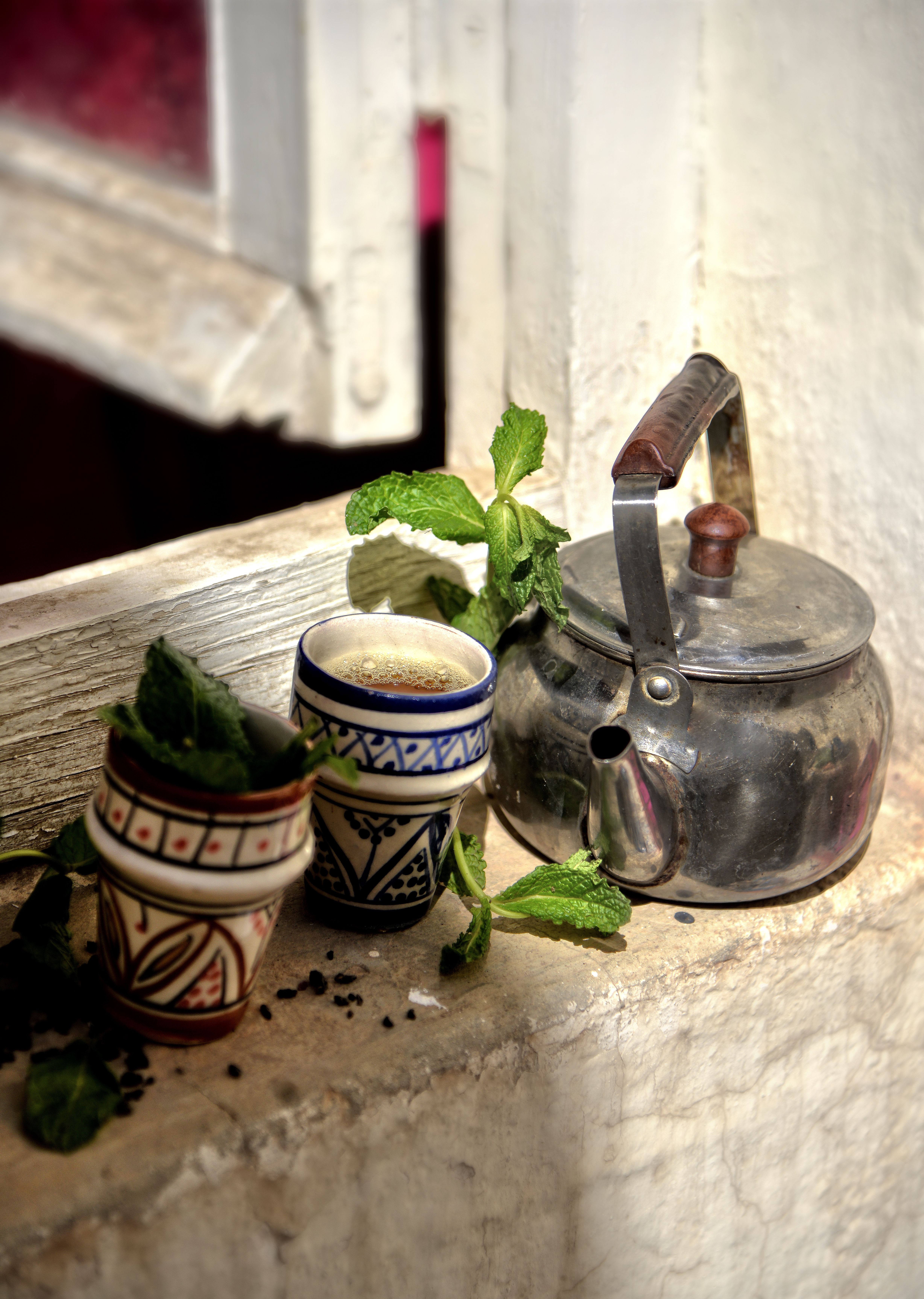 Théière en métal, deux petits verres peints et feuilles de menthe fraîche posés sur un rebord de fenêtre passé à la chaux