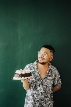 A joyful man in a patterned shirt holds a chocolate cake against a dark green background.