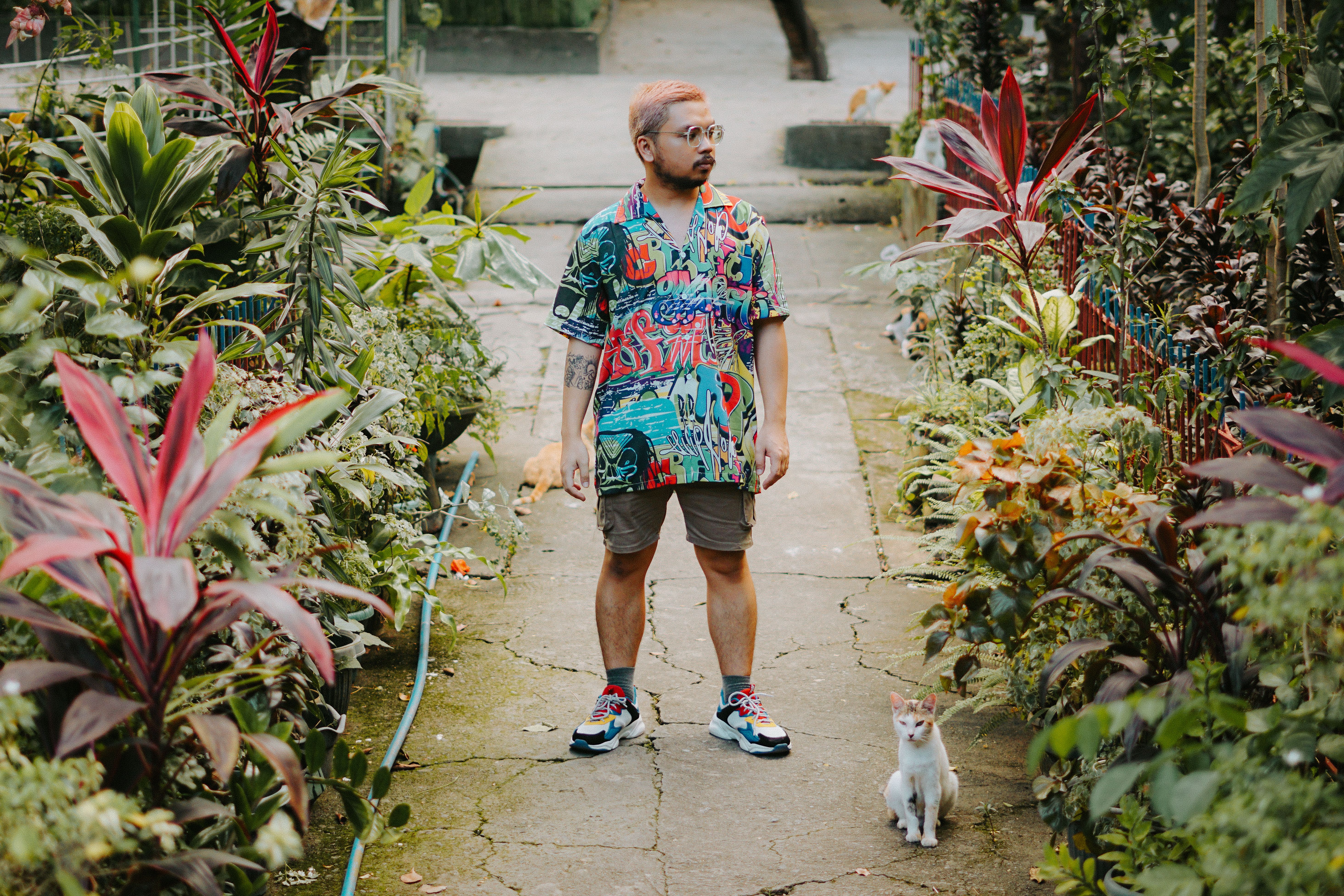 A man stands confidently in a vibrant shirt surrounded by lush greenery and tropical plants.