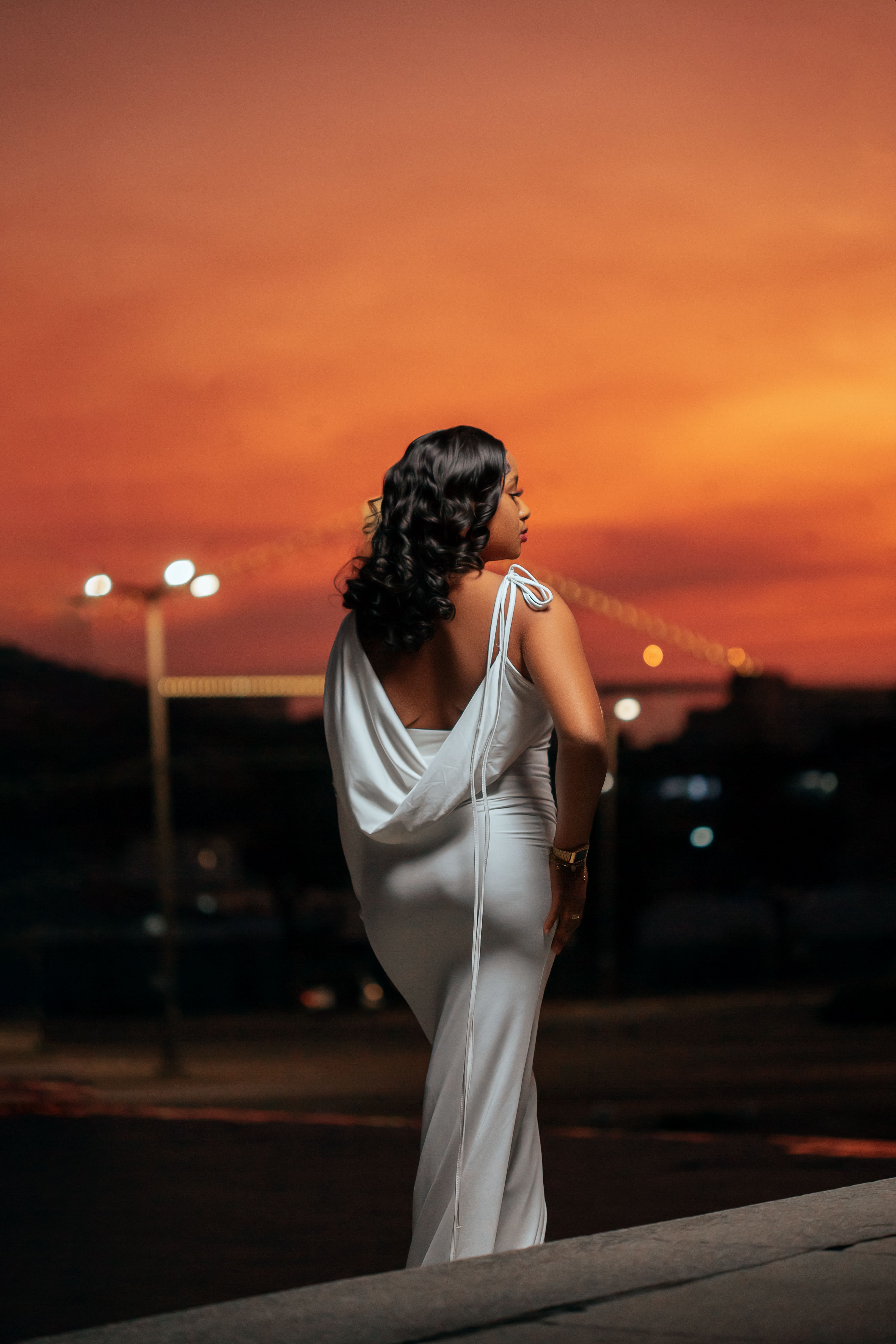 A woman in an elegant white dress stands against a vibrant sunset backdrop, exuding grace.