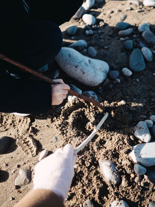 Free Two people using sticks to draw in the sand on a pebble beach, with rocks scattered around. Stock Photo